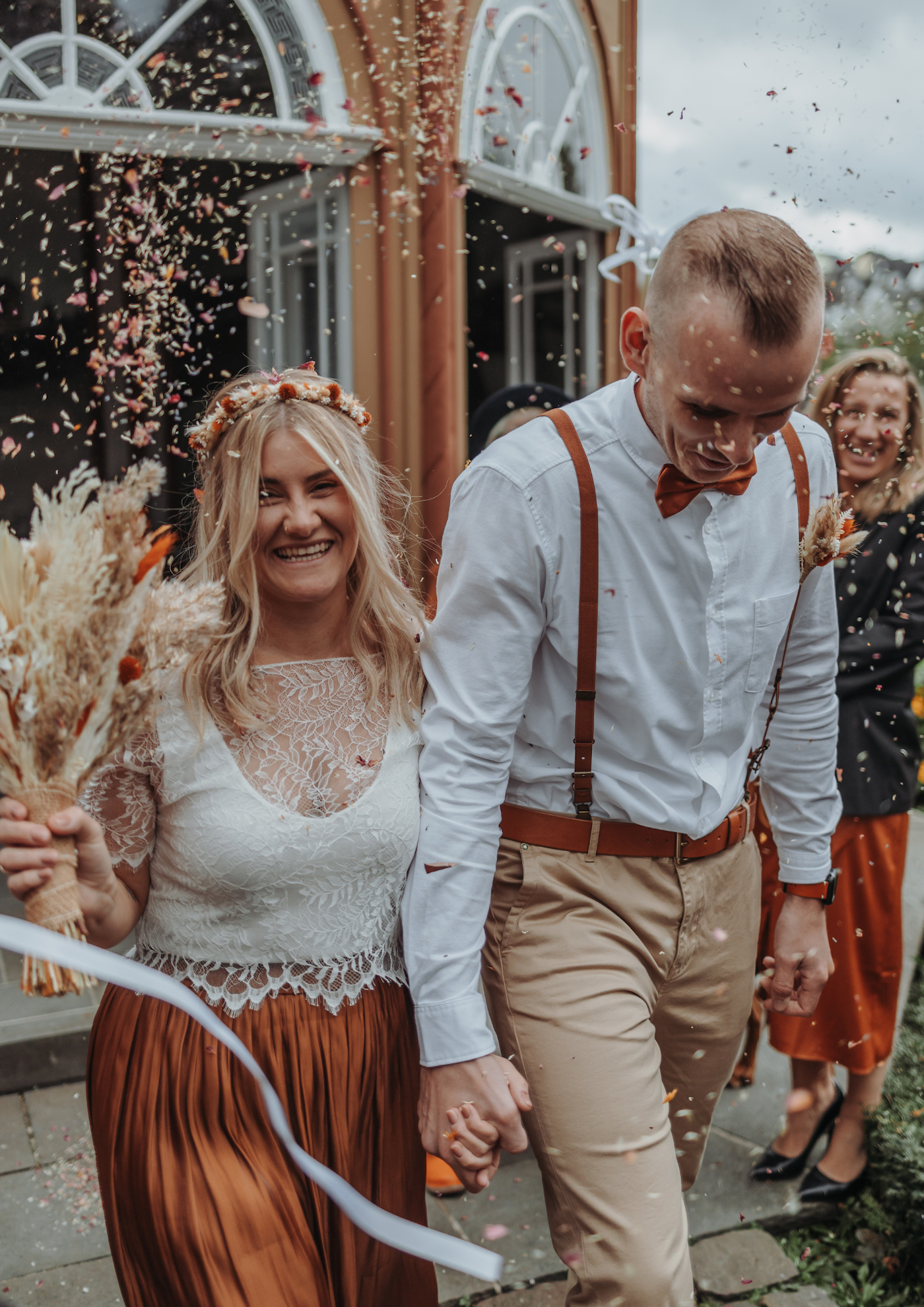 Hochzeitstag von Brautpaar strahelnd mit Brautstrauss und Bluetenkonfetti